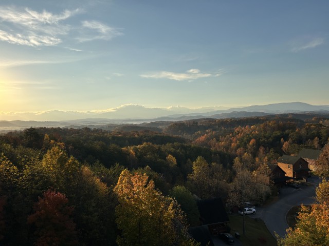 A bright forest in fall with yellow and red leaves from above. The sky is bright blue with a shining sun and mountain ranges extending far into the horizon.